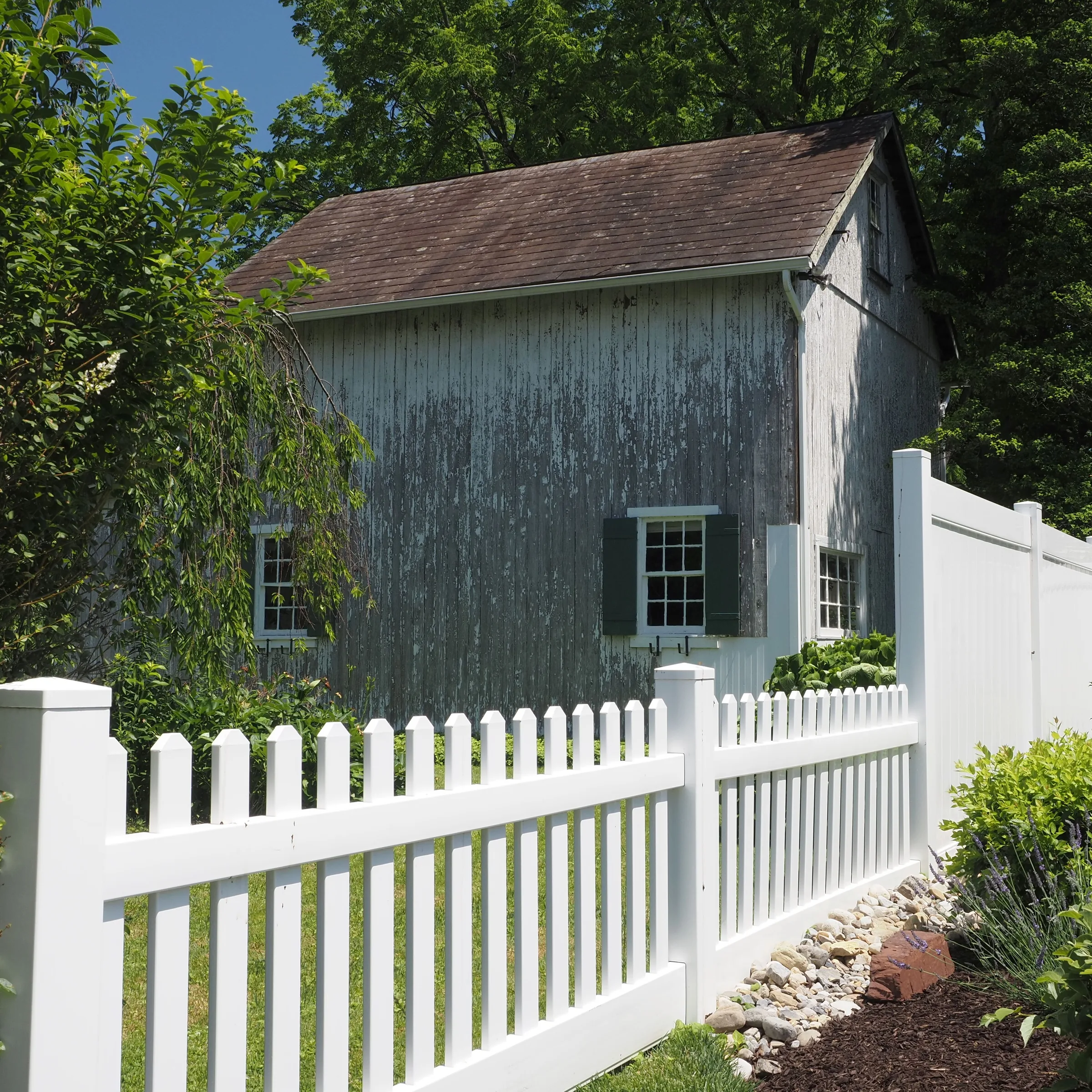 Weathered gray cottage with white picket fence and green shutters surrounded by trees.